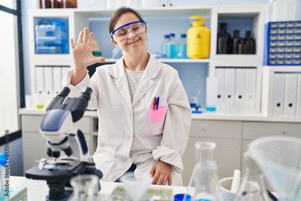 Hispanic girl with down syndrome working at scientist laboratory showing and pointing up with fingers number five while smiling confident and happy.