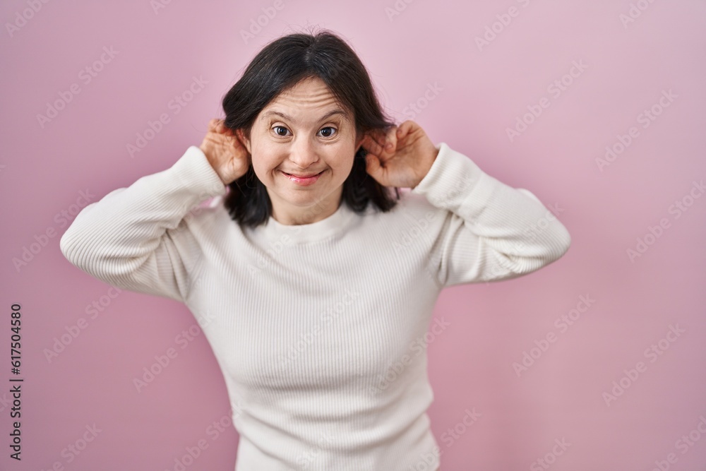 Woman with down syndrome standing over pink background smiling pulling ears with fingers, funny gesture. audition problem Woman with down syndrome standing over pink background smiling pulling ears with fingers, funny gesture. audition problem
