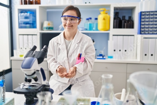 Hispanic girl with down syndrome working at scientist laboratory with hands together and crossed fingers smiling relaxed and cheerful. success and optimistic