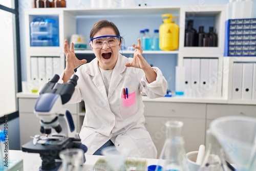 Hispanic girl with down syndrome working at scientist laboratory shouting frustrated with rage  hands trying to strangle  yelling mad