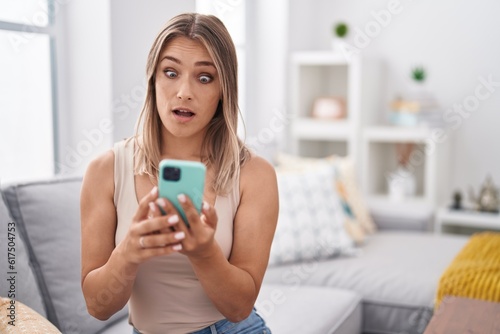 Blonde caucasian woman using smartphone typing message sitting on the sofa scared and amazed with open mouth for surprise, disbelief face