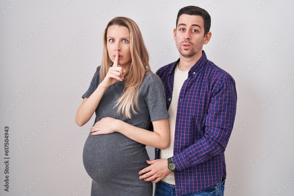 Young couple expecting a baby standing over white background asking to be quiet with finger on lips. silence and secret concept. Young couple expecting a baby standing over white background asking to be quiet with finger on lips. silence and secret concept.