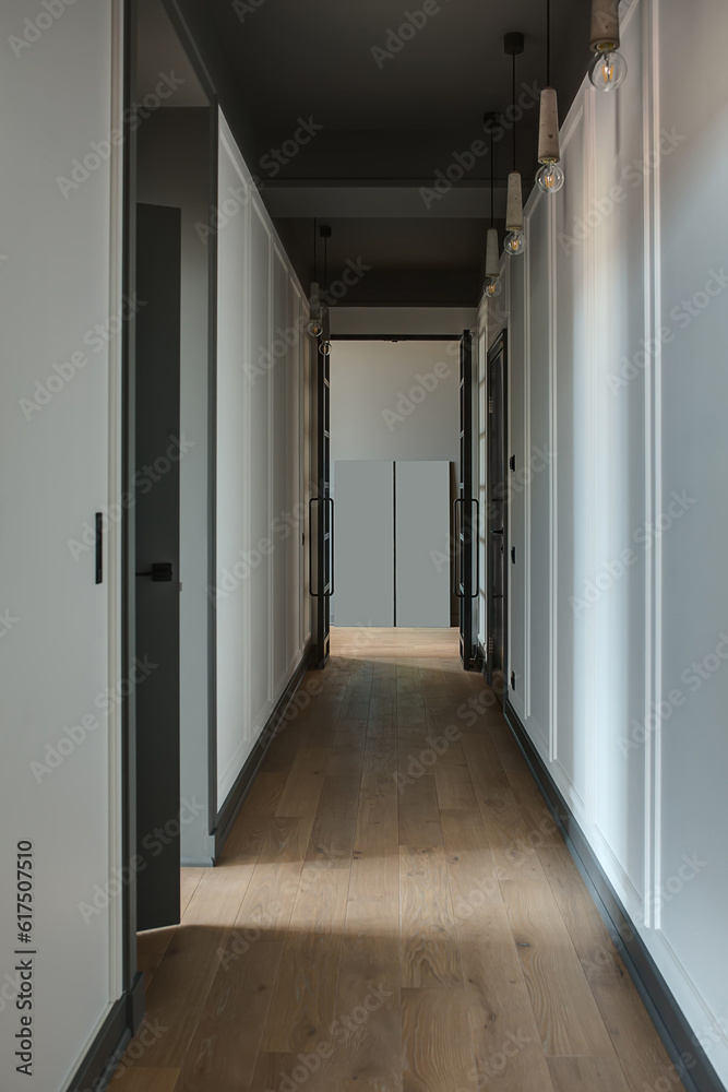 Modern style corridor with white walls and a parquet on the floor. There are black doors and hanging lamps on the ceiling. Vertical. Modern style corridor with white walls and a parquet on the floor. There are black doors and hanging lamps on the ceiling. Vertical.