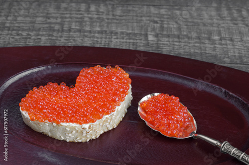 Sandwich with red caviar in a heart shape and spoon on wooden tray and grey background. Analog products.