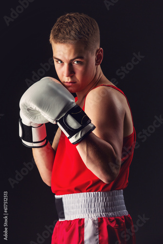 Young handsome boxer sportsman in red boxer suit and white gloves standing on black backgound. Copy space.