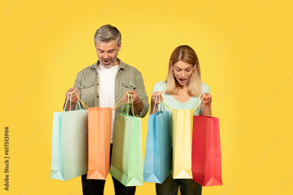 Great sales concept. European spouses holding and looking inside open shopper bags, standing on yellow background Great sales concept. European spouses holding and looking inside open shopper bags, standing on yellow background