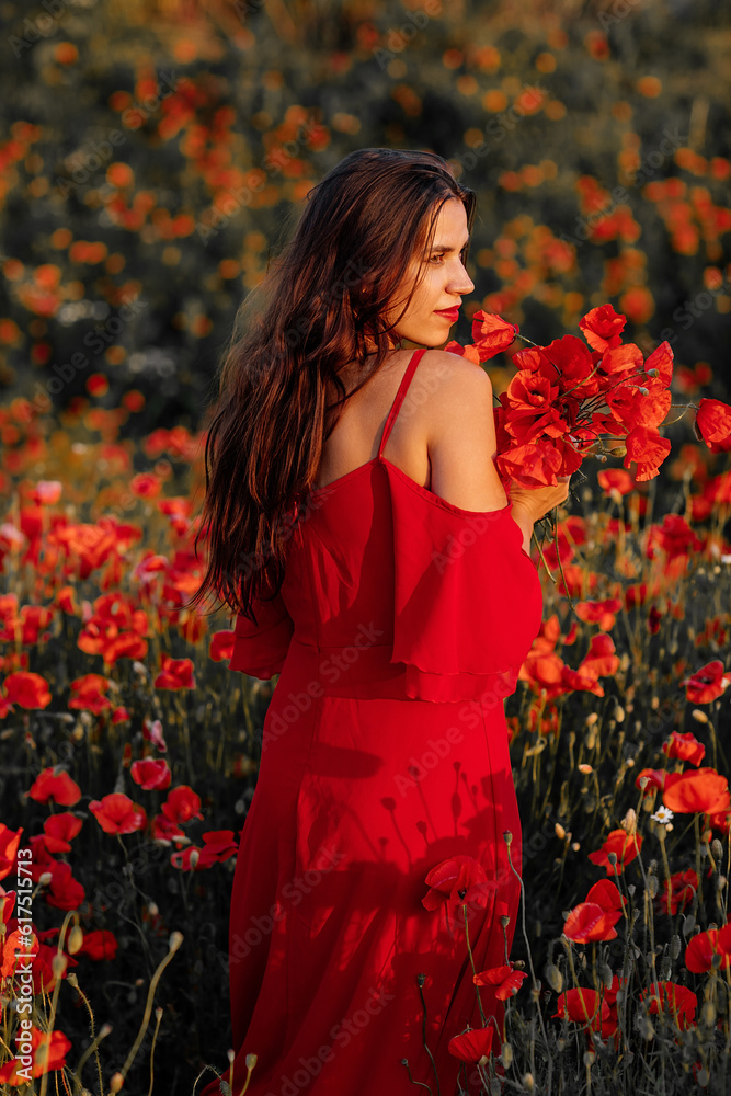 Naklejka premium Beautiful young woman with brown hair in red long dress in field with poppies in evening at sunset and holds poppy in hand, Summer countryside nature flowers, Female relaxing, Rural simple life