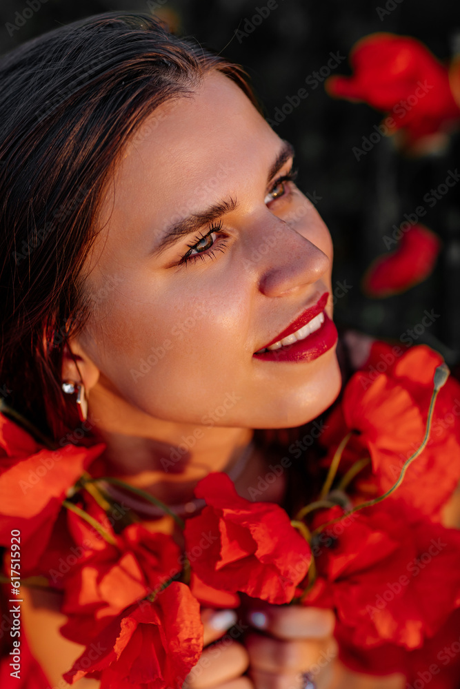 Naklejka premium Beautiful young woman with brown hair in red long dress in field with poppies in evening at sunset and holds poppy in hand, Summer countryside nature flowers, Female relaxing, Rural simple life