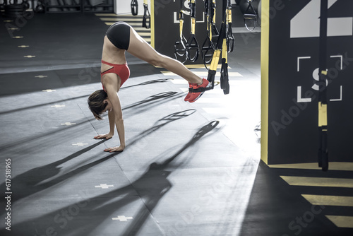 Amazing girl stands on the hands upside down on the floor in the gym. Her feet are on the TRX straps. She wears red top and sneakers, black shorts. Horizontal.