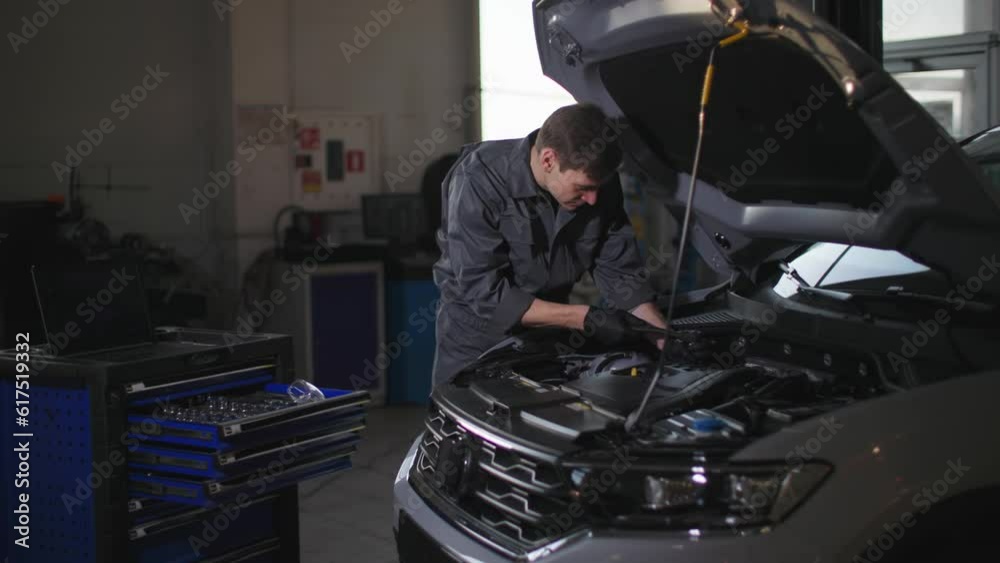 service vehicle, young male mechanic in uniform using a tool checks the engine under the hood of a car at a technical station service vehicle, young male mechanic in uniform using a tool checks the engine under the hood of a car at a technical station