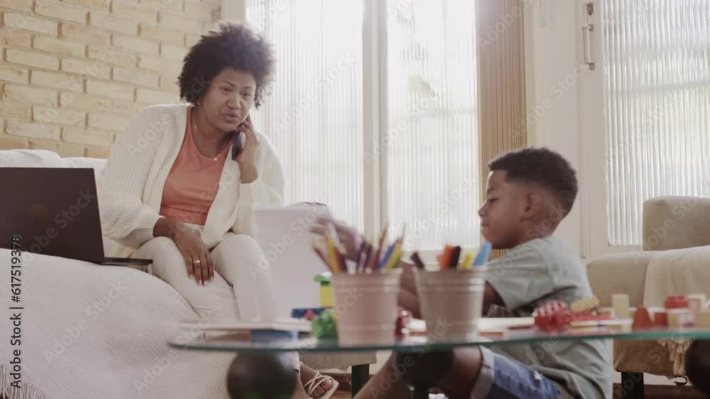 mulher Brasileira falando no celular e usando o computador no sofa da sala para resolver questoes do trabalho enquanto seu faz a licao de casa brinca com seu filho sentado no chao