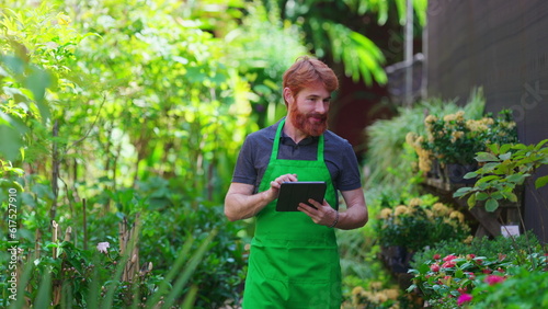 A happy male florist inspecting plant inventory at retail store with tablet. Young man wearing green apron walking toward camera and smiling at camera