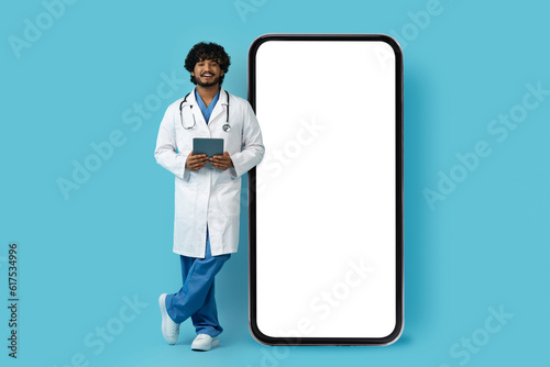 Friendly eastern doctor with digital pad standing by big phone