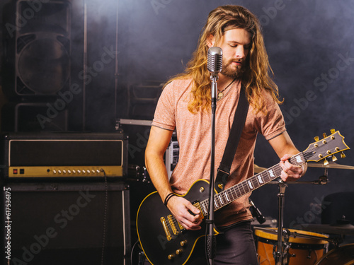Photo of a young man with long hair and beard playing electric guitar on stage.