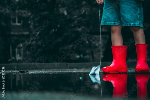 child in red rubber boots holds paper ships in his hands