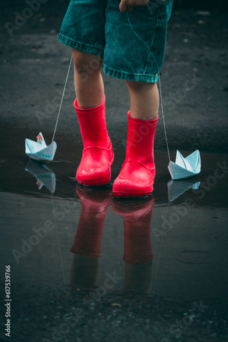 child in red rubber boots holds paper ships in his hands