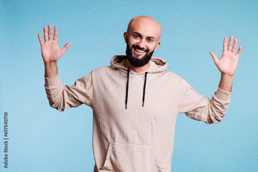 Fototapeta premium Smiling arab man with cheerful facial expression raising hands studio portrait. Young bearded arabian person posing, showing palms and looking at camera with joyful emotions