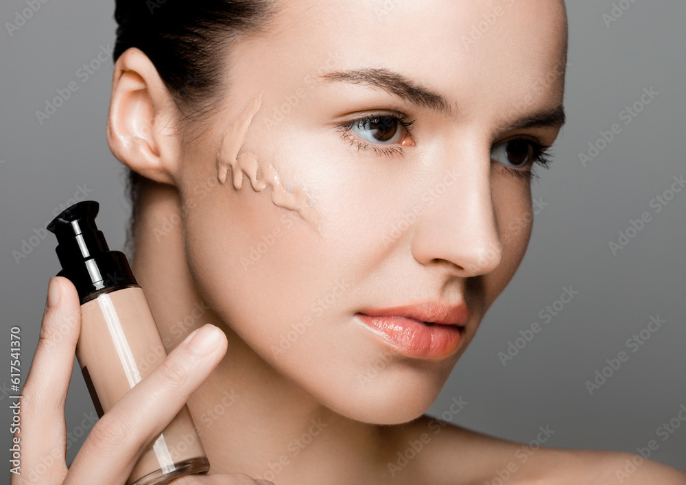 Beauty fashion model girl holding foundation tube container with foundation on her cheek on grey background