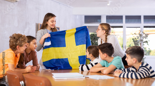Female teacher tells her classmates about the country of Sweden, holding a flag in her hands