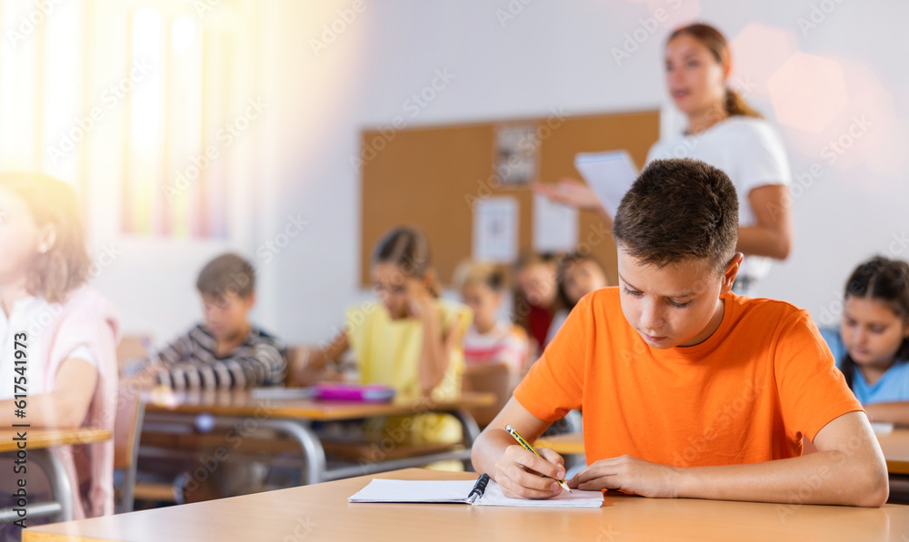 Fototapeta premium Portrait of focused preteen schoolboy writing exercises in workbook during lesson in classroom ..