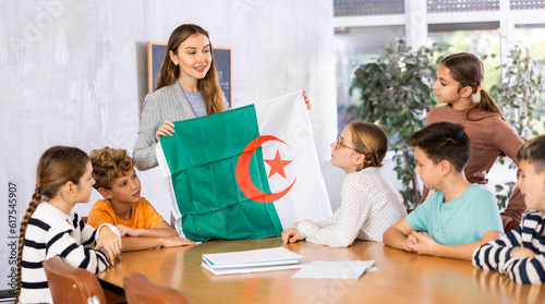 Smiling young woman teacher showing state flag of Algeria and telling preteens schoolchildren history of country during lesson in class
