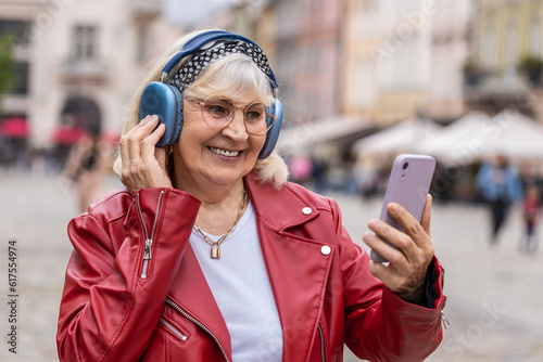 Happy relaxed overjoyed senior woman in wireless headphones choosing, listening favorite energetic disco rock n roll music in smartphone dancing outdoors. Grandmother walking in city street outside
