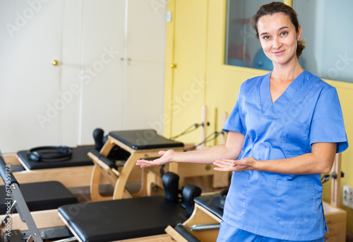Smiling professional female physiotherapist polite inviting to rehab center with pilates equipment. Concept of modern approach to physical medicine, physiotherapy exercises and rehabilitation
