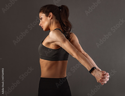 Sporty muscular woman with ponytail doing stretching workout the shoulders, blades and arms in sport bra, holding the hands back in palm lock behind on dark grey background