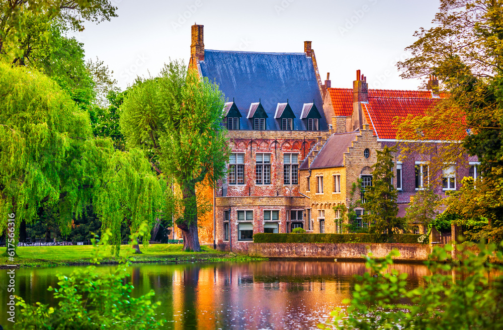 Fototapeta premium Vintage building over lake of love in Minnewater park in Bruges Belgium near Beguinage monastery of Beguines. Picturesque landscape with green trees sunset time.