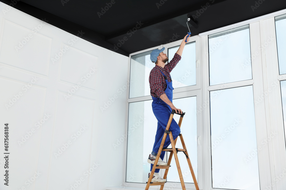 Worker in uniform painting ceiling with roller on stepladder indoors