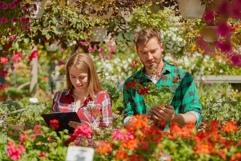 Male gardener standing with potted flower while female coworker doing paperwork in the garden.
