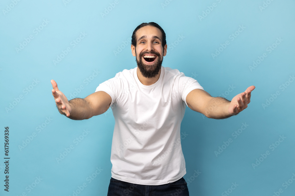 Welcome, wide open hug. Portrait of man with beard wearing white T-shirt stretching hands to camera and smiling broadly, going to embrace, share love. Indoor studio shot isolated on blue background. Welcome, wide open hug. Portrait of man with beard wearing white T-shirt stretching hands to camera and smiling broadly, going to embrace, share love. Indoor studio shot isolated on blue background.