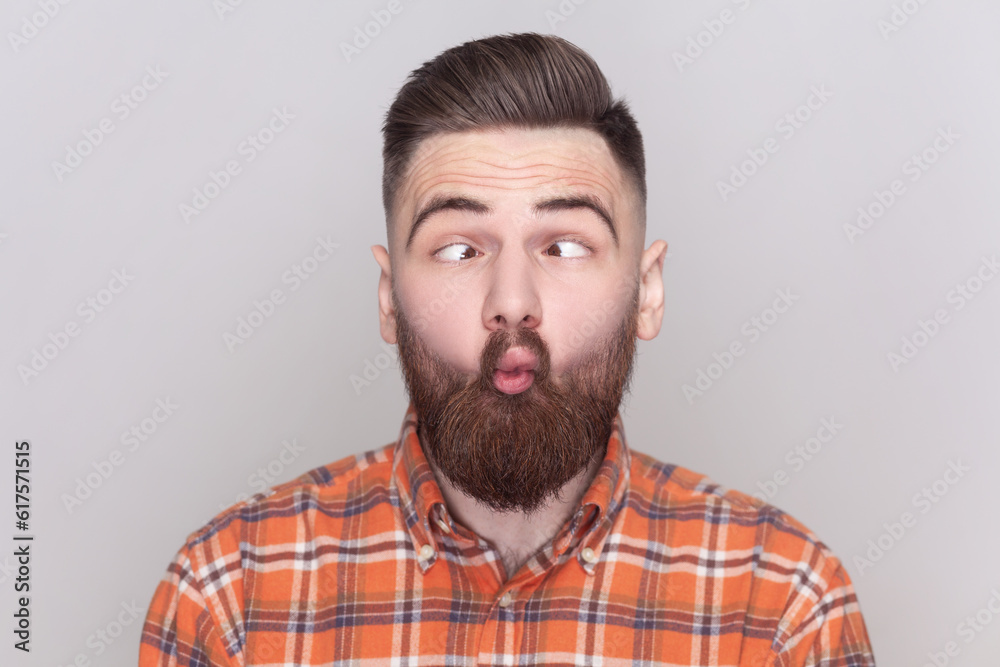 Obraz premium Portrait of funny crazy bearded man standing, crossed eyes with fish lips gesture, fool around, having fun, wearing checkered shirt. Indoor studio shot isolated on gray background.