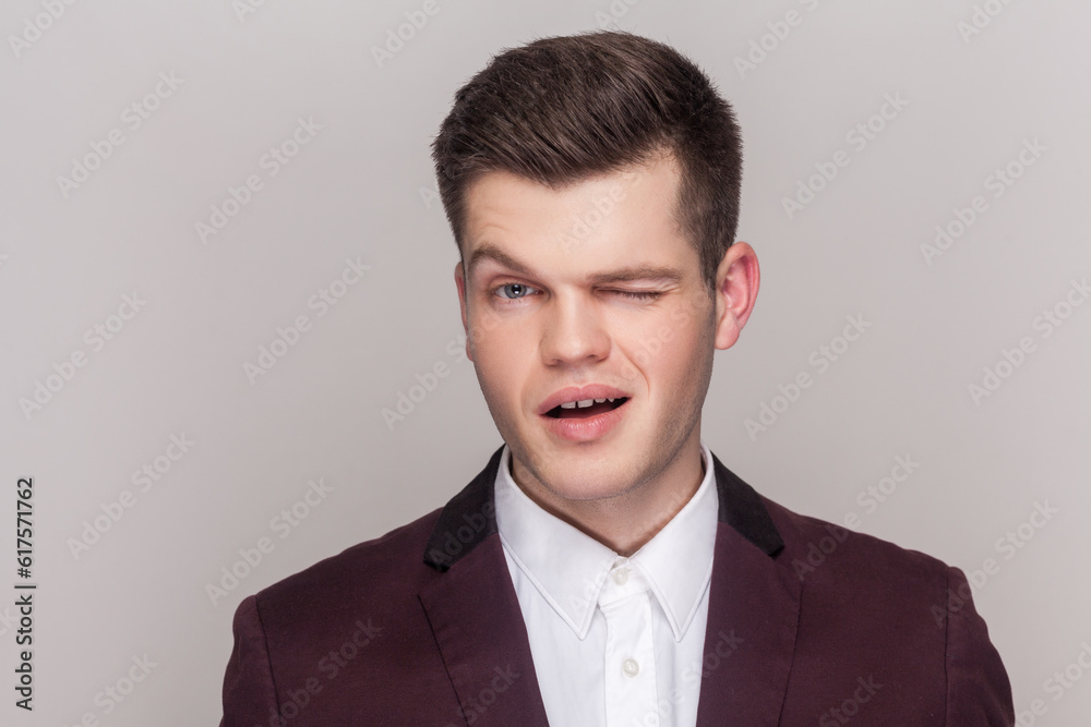 Portrait of playful handsome young man has fun and winks eye, smiles with teeth makes funny grimace, wearing violet suit and white shirt. Indoor studio shot isolated on grey background. Portrait of playful handsome young man has fun and winks eye, smiles with teeth makes funny grimace, wearing violet suit and white shirt. Indoor studio shot isolated on grey background.