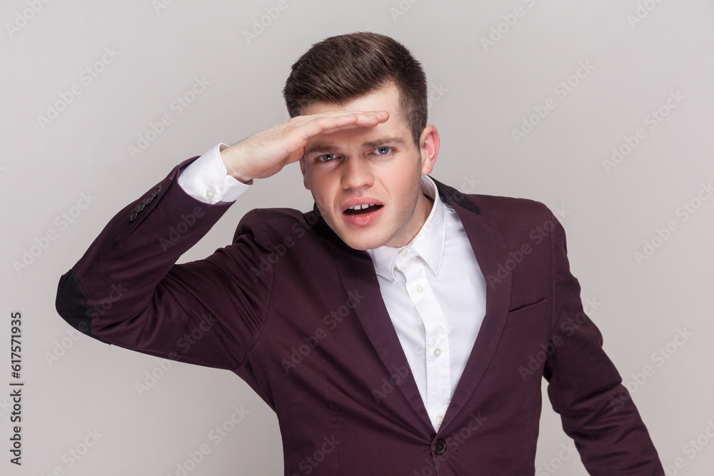 Man peering into distance, looking far away with attentive view, searching on horizon and curious to discover, wearing violet suit and white shirt. Indoor studio shot isolated on grey background. Man peering into distance, looking far away with attentive view, searching on horizon and curious to discover, wearing violet suit and white shirt. Indoor studio shot isolated on grey background.