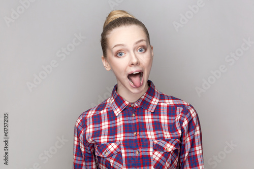 Portrait of foolish crazy playful woman with bun hairstyle looking at camera with open mouth and showing tongue out, wearing checkered shirt. Indoor studio shot isolated on gray background.