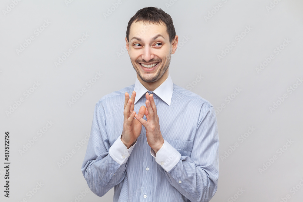 Portrait of cunning sly young adult man standing looking away with toothy smile, having devil plans, planning prank, wearing light blue shirt. Indoor studio shot isolated on gray background. - obrazy, fototapety, plakaty 