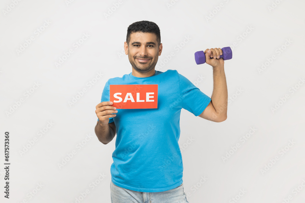 Portrait of smiling delighted man wearing blue T- shirt standing holding dumbbell and card with sale inscription, discounts for fitness season ticket. Indoor studio shot isolated on gray background Portrait of smiling delighted man wearing blue T- shirt standing holding dumbbell and card with sale inscription, discounts for fitness season ticket. Indoor studio shot isolated on gray background