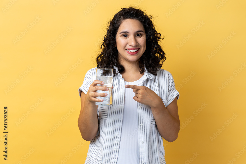 Portrait of smiling happy positive beautiful woman with dark wavy hair standing with glass of water, pointing at beverage with finger. Indoor studio shot isolated on yellow background. Portrait of smiling happy positive beautiful woman with dark wavy hair standing with glass of water, pointing at beverage with finger. Indoor studio shot isolated on yellow background.