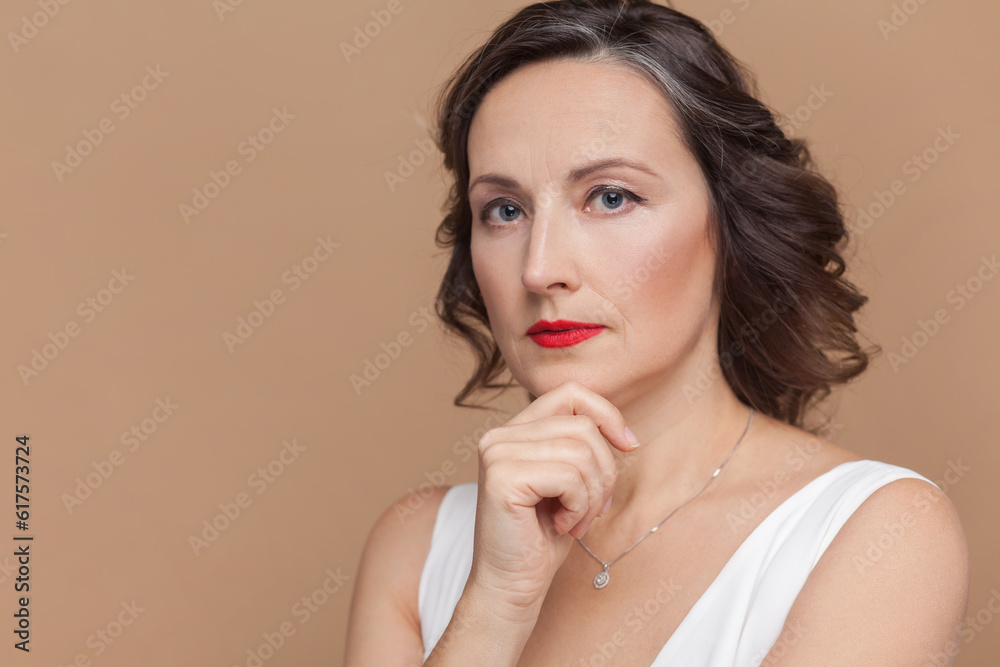Closeup of serious pensive attractive beautiful middle aged woman with wavy hair and makeup, looking at camera, holding chin, wearing white dress. Indoor studio shot isolated on light brown background Closeup of serious pensive attractive beautiful middle aged woman with wavy hair and makeup, looking at camera, holding chin, wearing white dress. Indoor studio shot isolated on light brown background