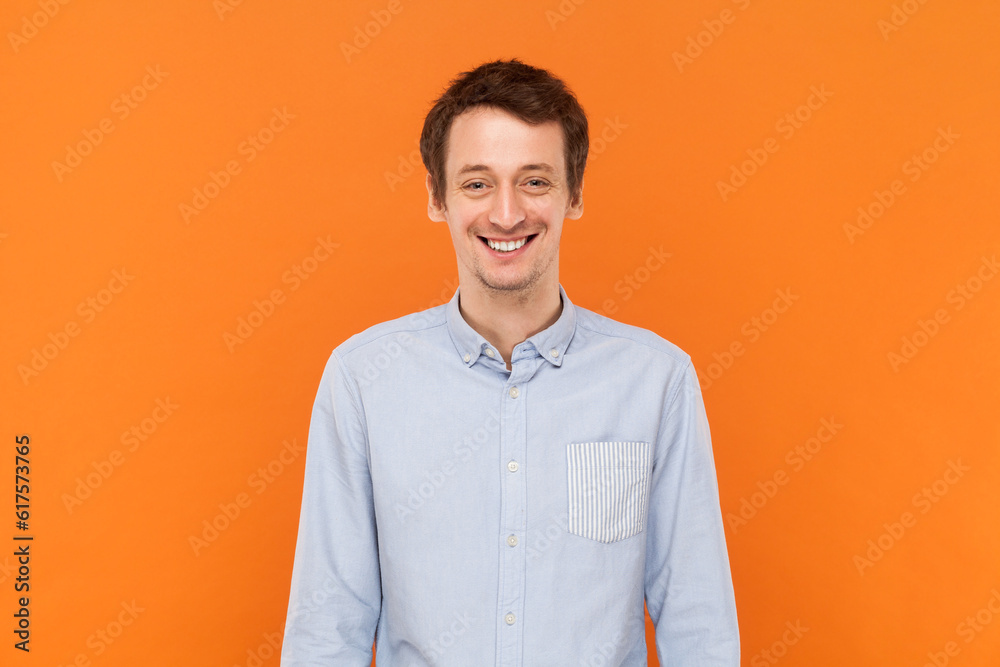 Portrait of overjoyed positive happy man standing looking at camera with toothy smile, expressing happiness, wearing light blue shirt. Indoor studio shot isolated on orange background. Portrait of overjoyed positive happy man standing looking at camera with toothy smile, expressing happiness, wearing light blue shirt. Indoor studio shot isolated on orange background.
