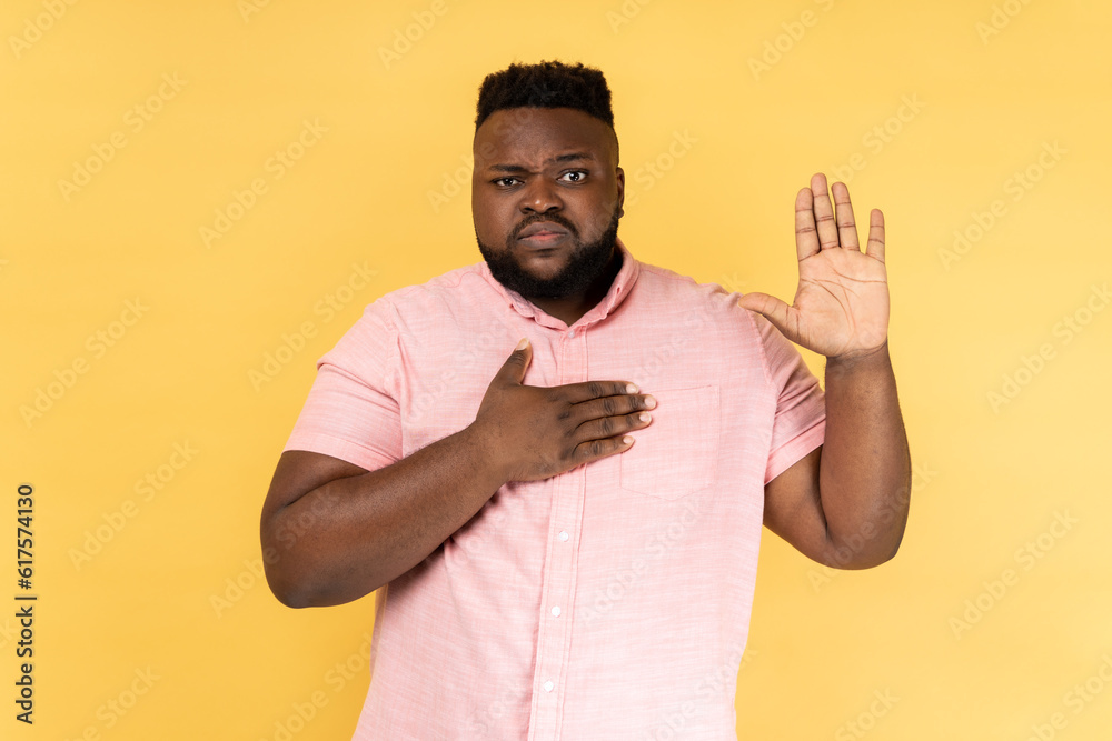 Portrait of serious patriotic man wearing pink shirt holding hand on heart, juryman swearing to speak truth in court, honor and conscience. Indoor studio shot isolated on yellow background. Portrait of serious patriotic man wearing pink shirt holding hand on heart, juryman swearing to speak truth in court, honor and conscience. Indoor studio shot isolated on yellow background.