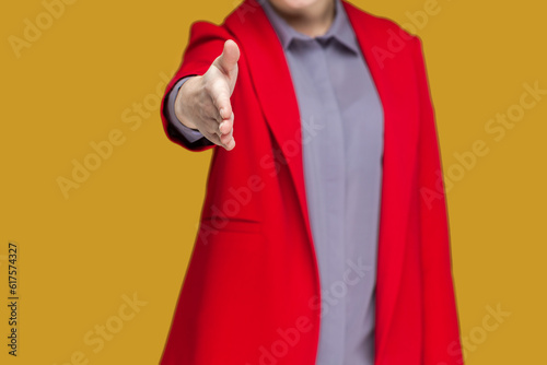 Closeup portrait of unknown woman standing with outstretched hand, saying hello or bye, handshaking, congratulating, wearing red jacket. Indoor studio shot isolated on yellow background.