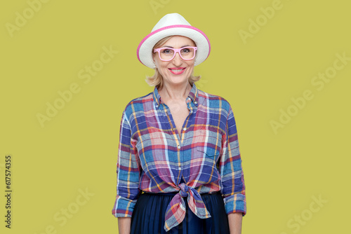 Portrait of smiling joyful senior woman wearing checkered shirt, hat and eyeglasses, looking at camera with toothy smile, expressing positive emotions. Indoor studio shot isolated on yellow background