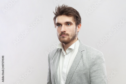Portrait of handsome attractive young bearded man standing with serious concentrated facial expression, looking away, wearing grey suit. Indoor studio shot isolated on gray background.
