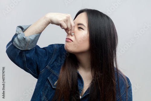 Portrait of disgusted brunette woman in blue denim jacket standing plugs nose as smells something stink and unpleasant, feels aversion. Indoor studio shot isolated on gray background.