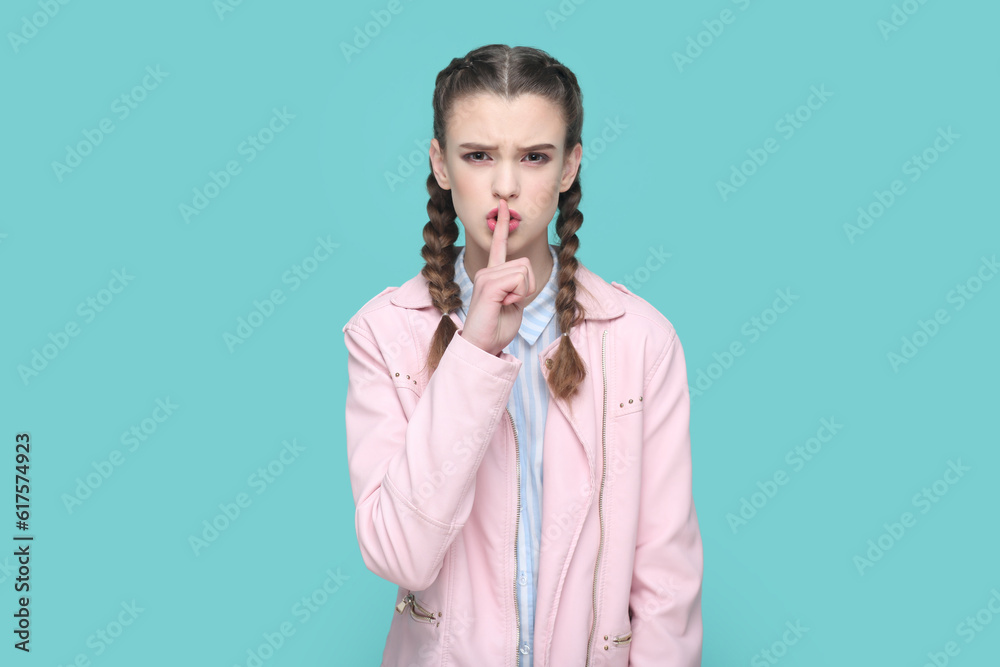 Portrait of serious teenager girl with braids wearing pink jacket keeps finger near lips, showing shh gesture, asking to keep silence. Indoor studio shot isolated on green background. Portrait of serious teenager girl with braids wearing pink jacket keeps finger near lips, showing shh gesture, asking to keep silence. Indoor studio shot isolated on green background.