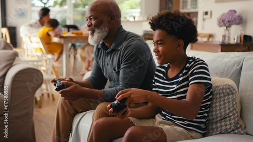 Grandfather and grandson sitting on sofa at home holding controllers playing video game together with multi-generation family in background - shot in slow motion