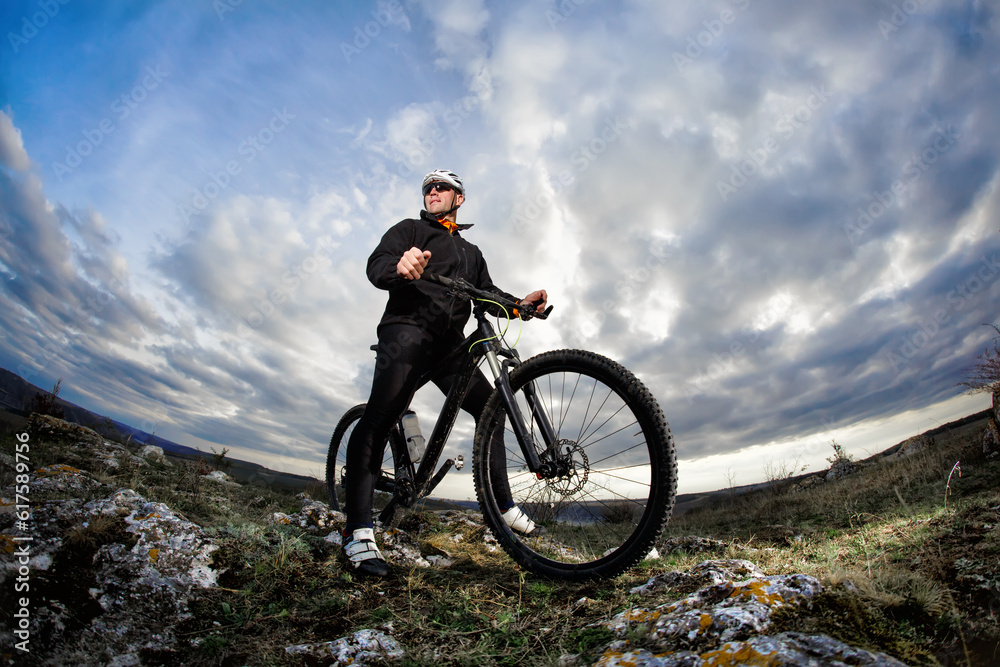 Photo below of the mountain cyclist in the black sportwear on the rocks against dramatic sky with clouds. Cyclist in the helmet and sunglasses. Beautiful landscape. Summer season in the coutryside. He