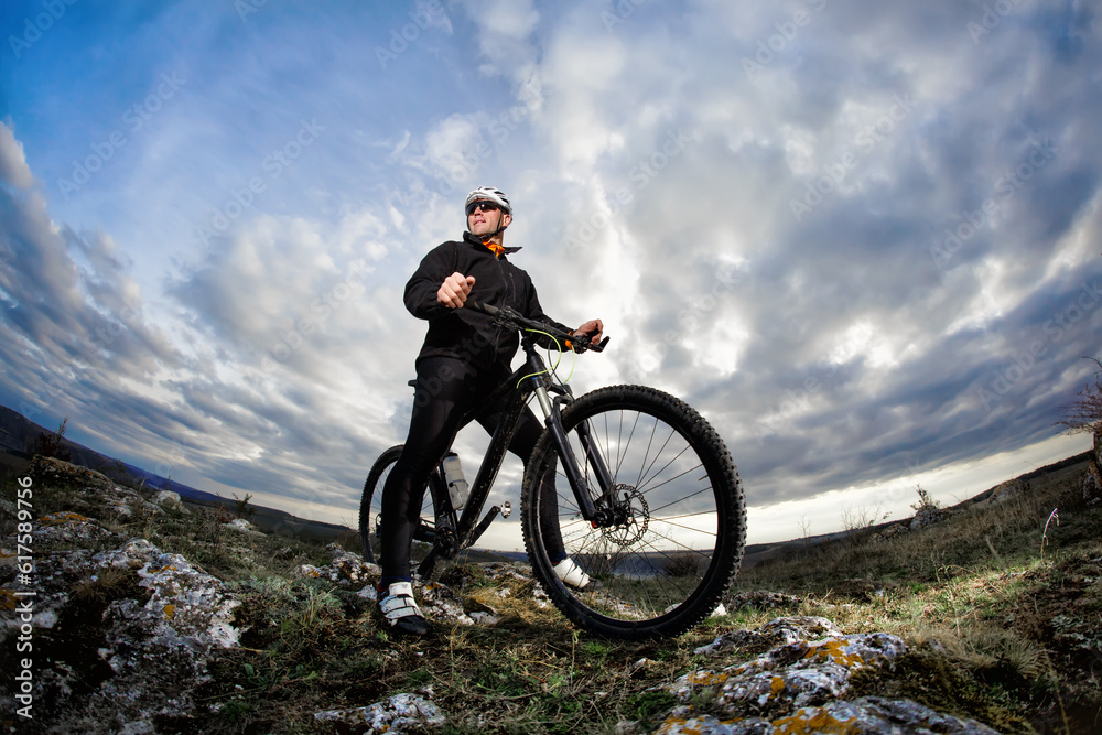 Naklejka premium Photo below of the mountain cyclist in the black sportwear on the rocks against dramatic sky with clouds. Cyclist in the helmet and sunglasses. Beautiful landscape. Summer season in the coutryside. He